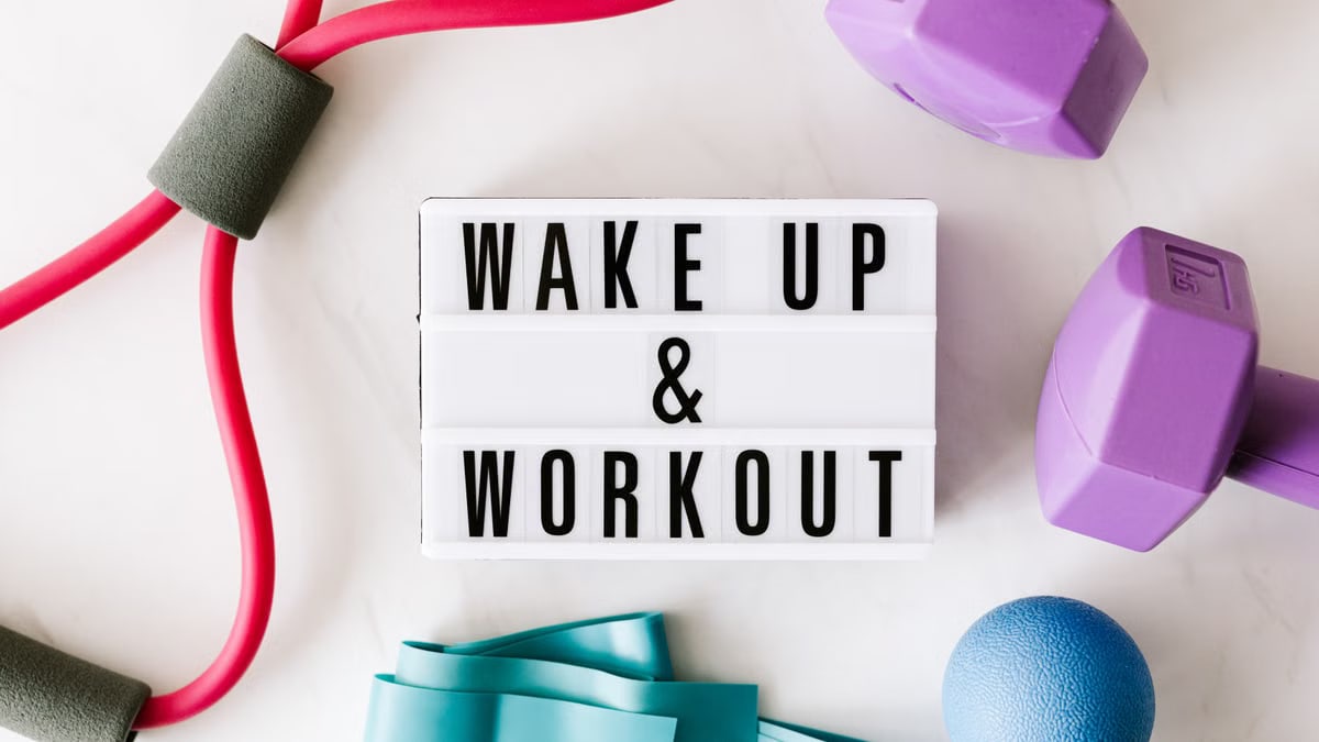 Top view closeup composition of wake up and workout inscription on light box surface among expanders and dumbbells and massage ball on white background